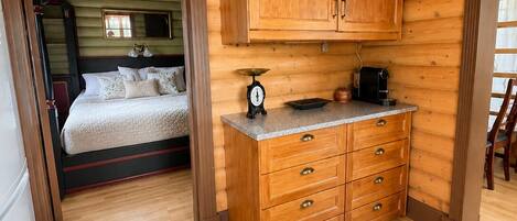 A wooden cabin interior with a bed, a kitchen area with cabinets and a scale, and a dining area with a chair.