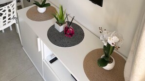 Uma área de jantar moderna com televisão de tela plana na parede, mesa de jantar branca, cadeiras e três plantas em vasos sobre jogos americanos de fibra natural.