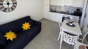 Uma sala de estar moderna com um sofá azul escuro, uma mesa de jantar branca com cadeiras e uma cozinha com armários e eletrodomésticos brancos.