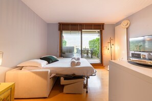 A bedroom with a bed, a TV, a desk, and a balcony with outdoor furniture. - Charmant studio au bord du lac avec sa terrasse et sa vue sur les montagnes (Duingt)