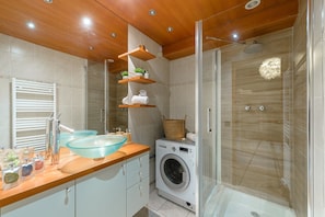 A modern bathroom with a glass shower enclosure, a built-in washing machine, and a wooden vanity with a glass bowl sink. - Charmant studio au bord du lac avec sa terrasse et sa vue sur les montagnes (Duingt)