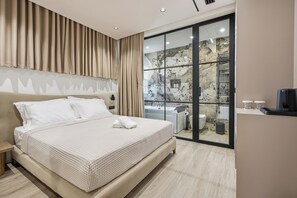A modern bedroom with a large bed, a sliding glass door to a bathroom, and a view of a stone wall. - Art Boutique Hotel Berat (Berat)