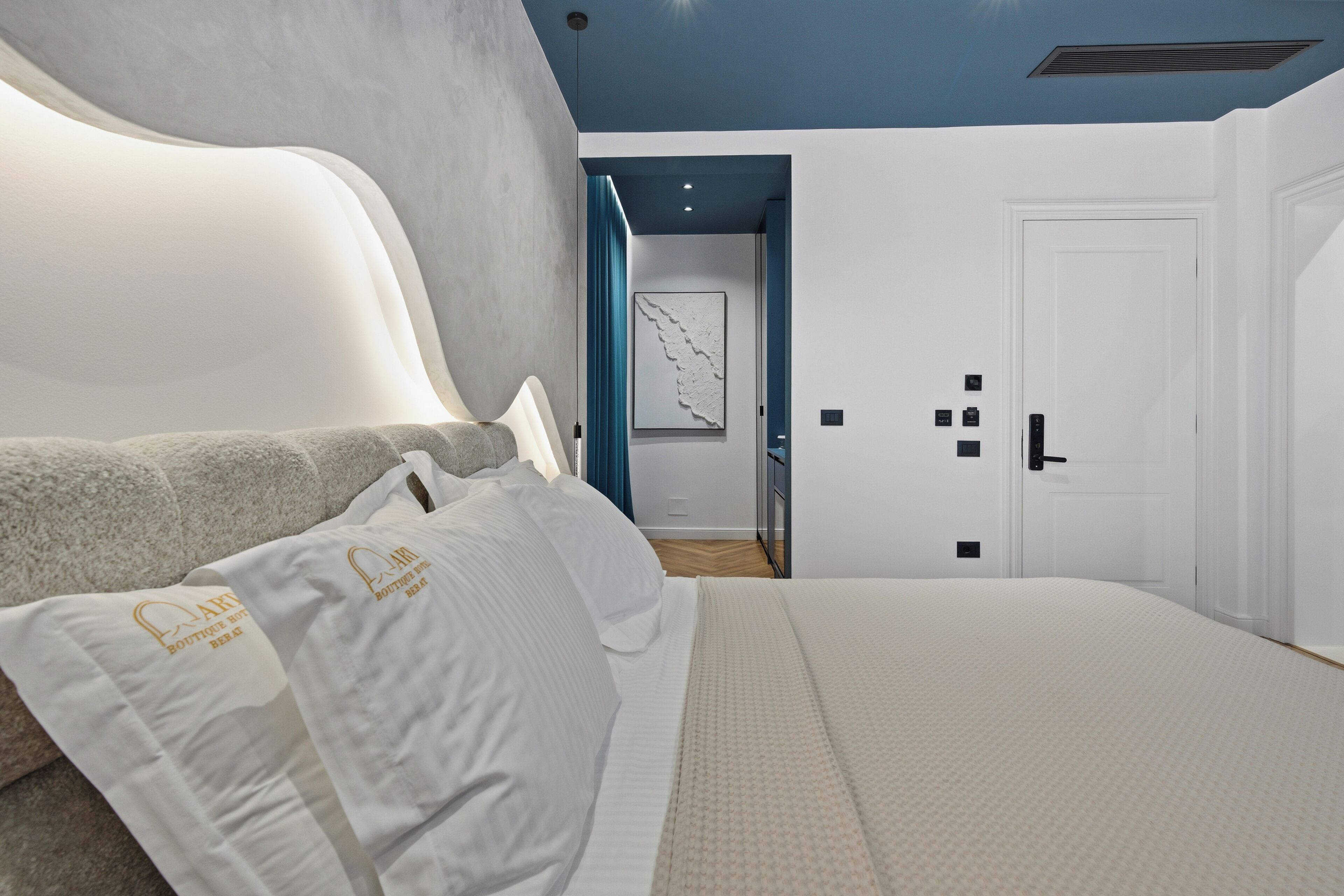 A modern bedroom with a large bed, a headboard, and a view of a hallway with a door and a framed picture.