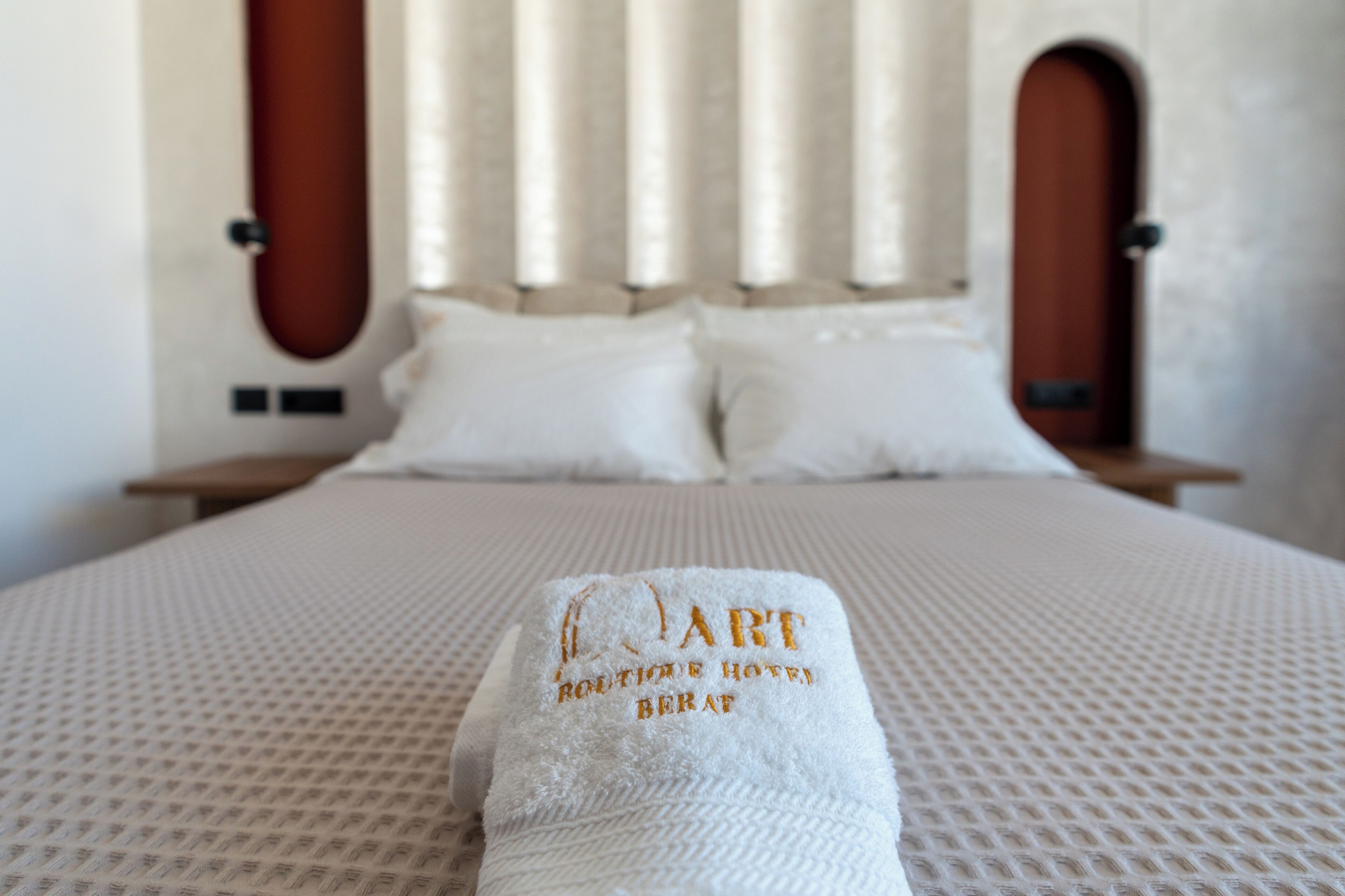 Ein ordentlich bezogenes Bett mit einem Handtuch mit dem Aufdruck 'ART Boutique Hotel', einem Kopfteil mit vertikalen Lamellen und zwei Nachttischen.