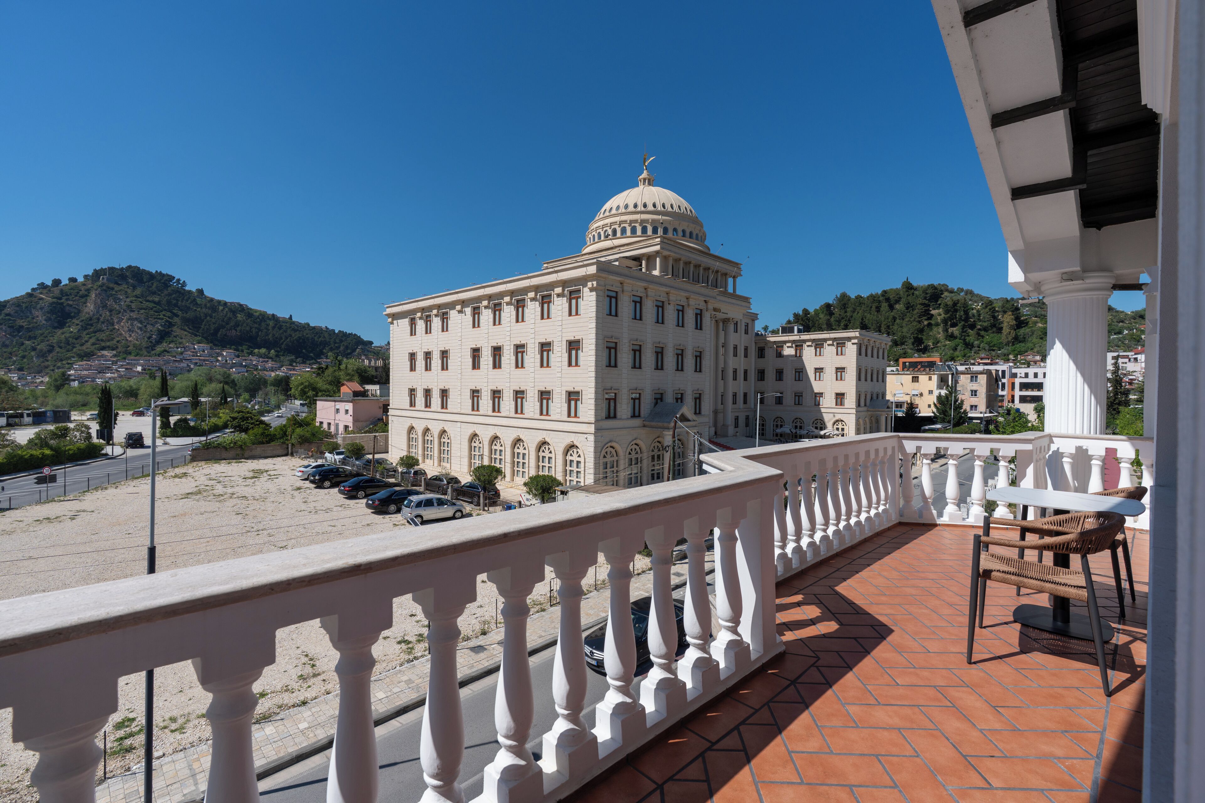 A building with a dome, a balcony with a white balustrade, and a view of a parking lot and hills in the background.