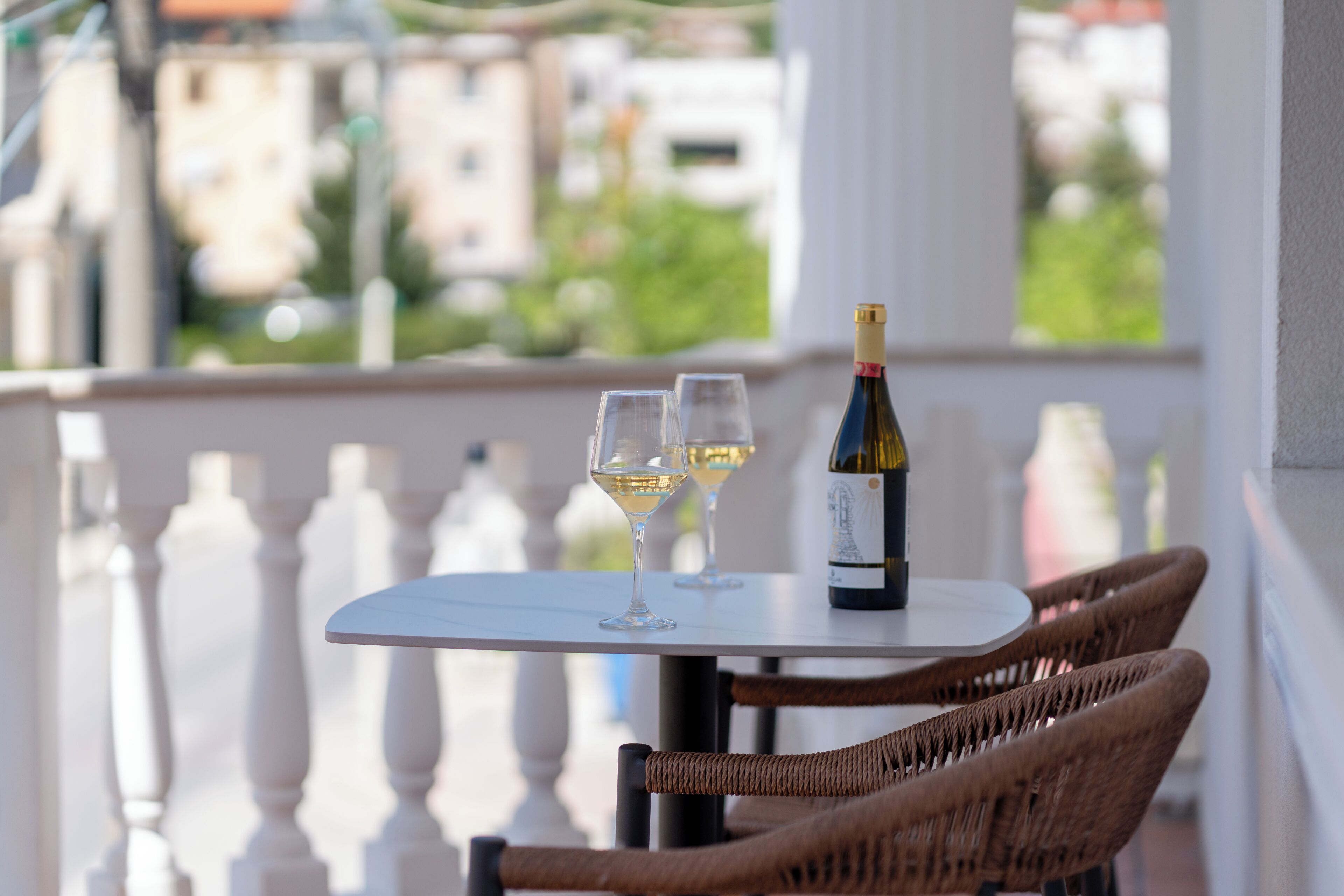A table with two wine glasses and a bottle on a balcony with wicker chairs and a railing.