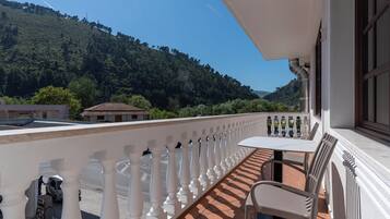 A balcony with a white balustrade, a table, and chairs, overlooking a hilly landscape with trees and a distant building.