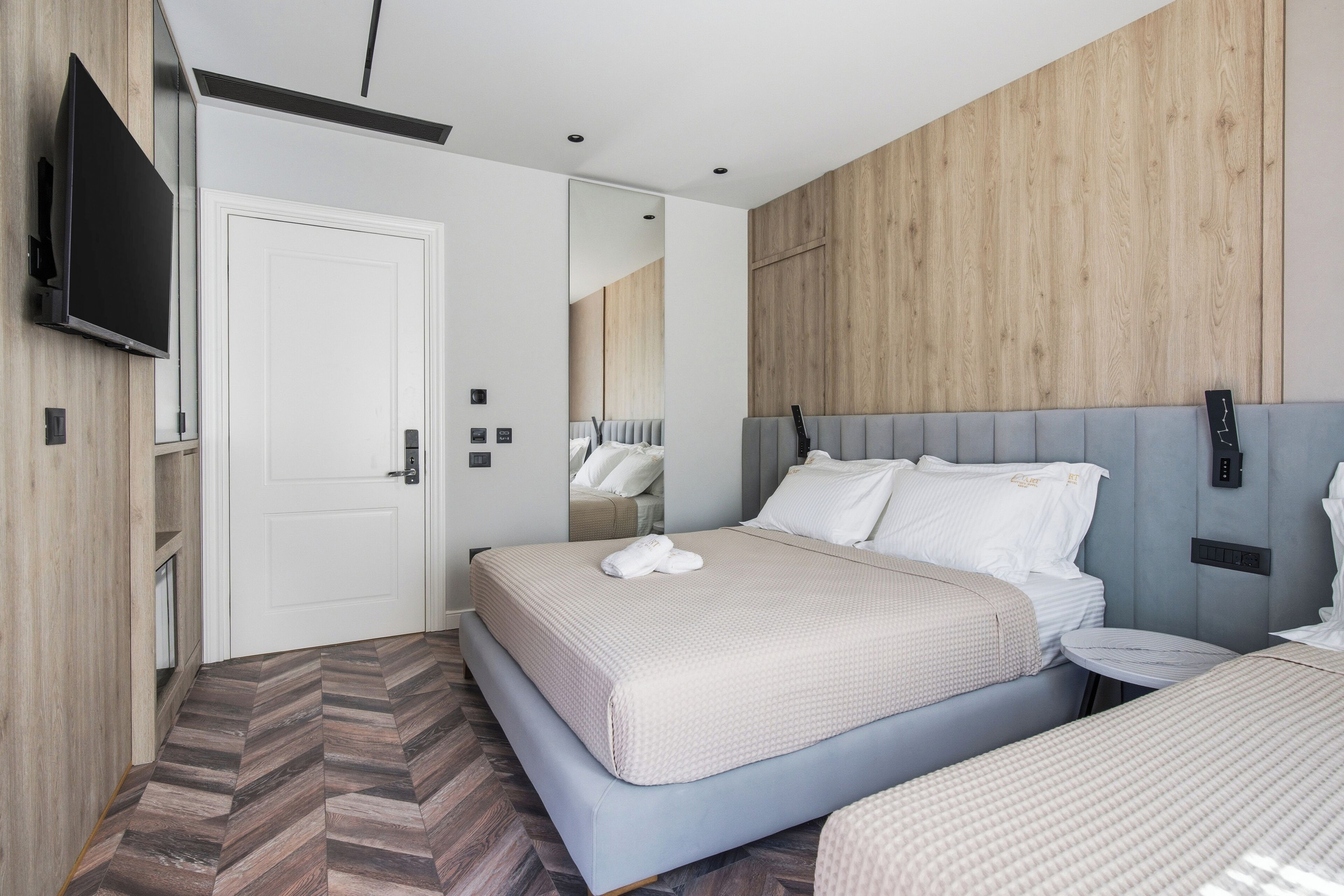 A modern hotel room with a large bed, a bedside table, a flat-screen TV mounted on the wall, and a wooden accent wall.