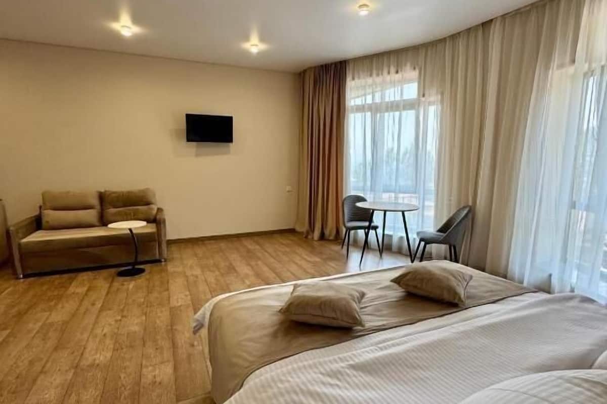 Una camera d'albergo con pavimento in legno, un divano, un letto con lenzuola bianche, un tavolino e una TV fissata al muro.