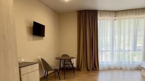 A spacious room with a wooden floor, a flat-screen TV mounted on the wall, a dining table with chairs, and a large window with curtains. - MARCO Hotel (Goris)