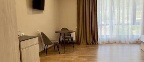 A spacious room with a wooden floor, a flat-screen TV mounted on the wall, a dining table with chairs, and a large window with curtains.