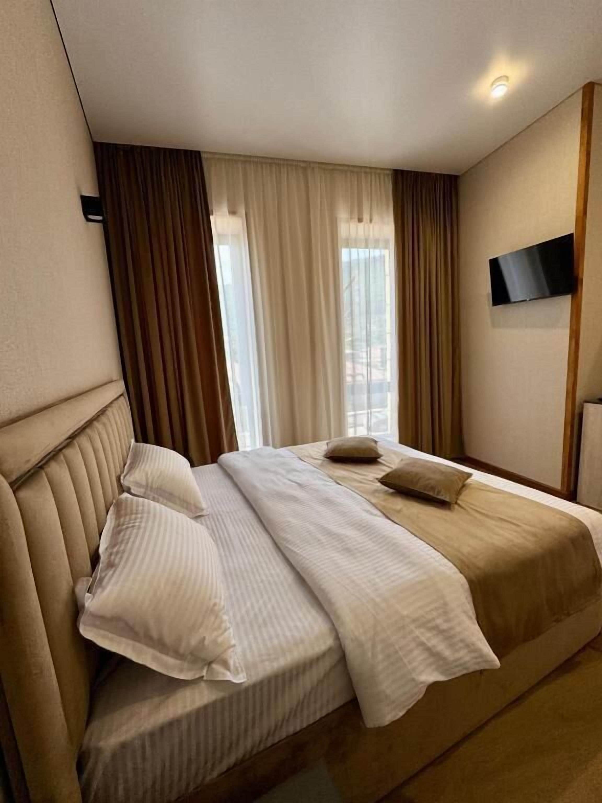A hotel room with a large bed, a flat-screen TV mounted on the wall, and a window with curtains.