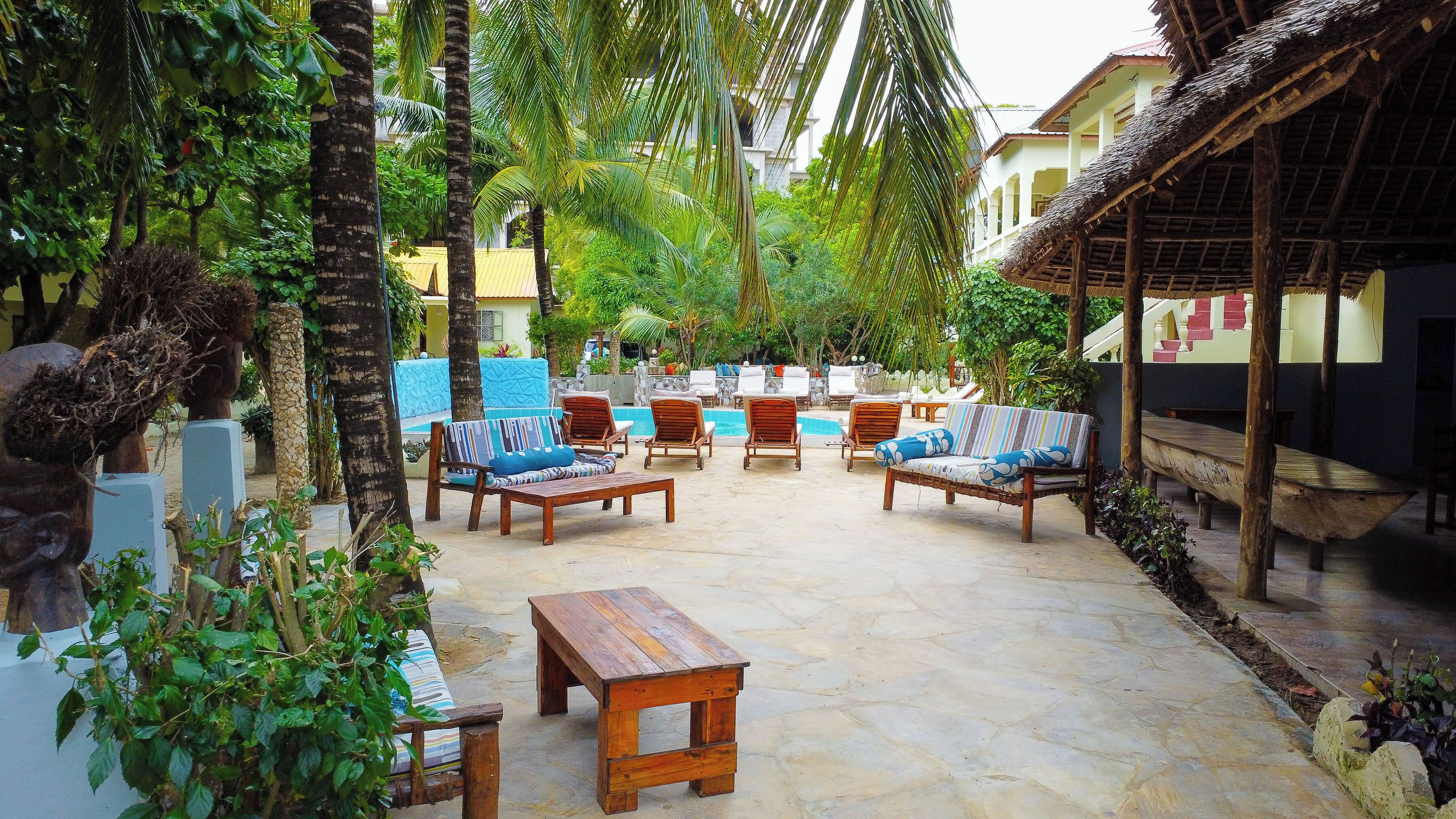 A tropical outdoor seating area with lounge chairs and a wooden table, surrounded by lush greenery and a pool.