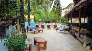 A tropical outdoor seating area with lounge chairs and a wooden table, surrounded by lush greenery and a pool.