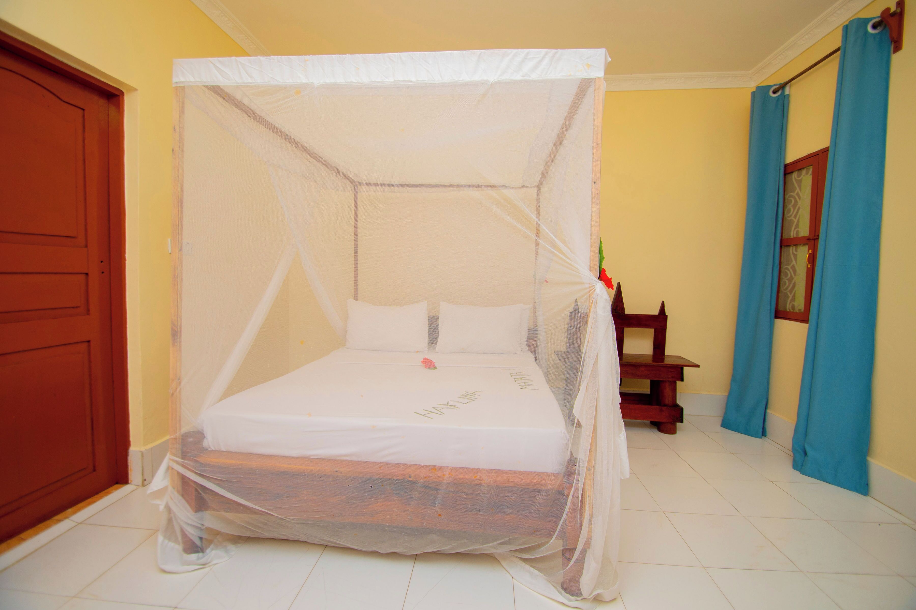Een bed met hemel, een houten stoel, een deur en een raam met gordijnen.