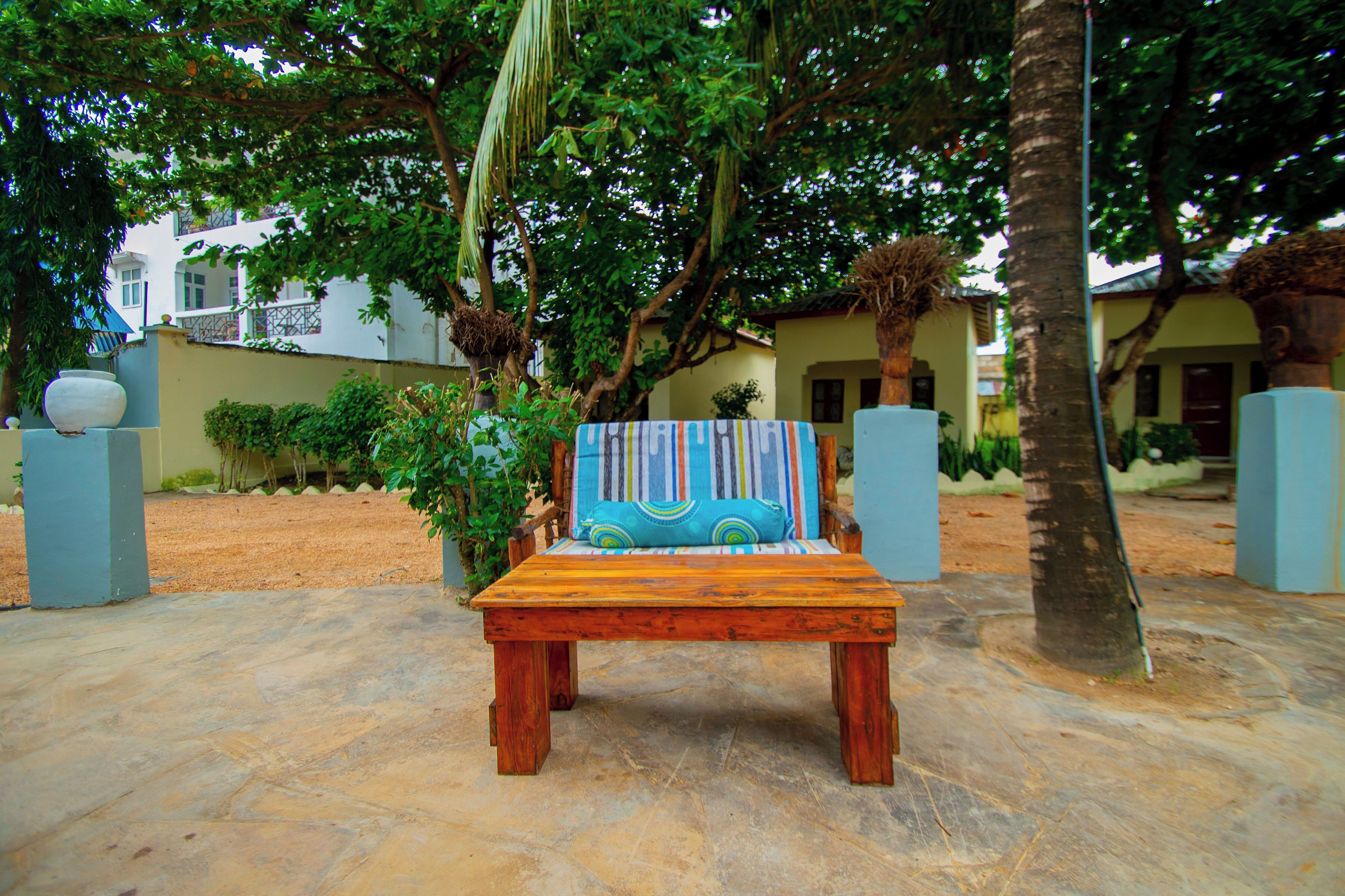 An outdoor seating area with a wooden bench and a colorful cushion, surrounded by trees and a building in the background.