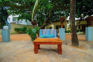 An outdoor seating area with a wooden bench and a colorful cushion, surrounded by trees and a building in the background. - Samridhi Bustani Oasis (Nungwi)