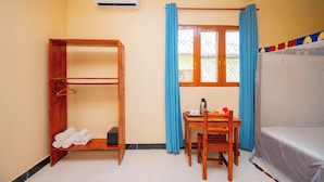 Quarto com uma cama de madeira estilo beliche, uma mesinha com chaleira, uma cadeira, uma estante de madeira com toalhas e uma janela com cortinas azuis.