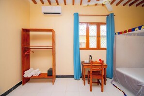 Una habitación con una cama litera de madera, una mesita con un hervidor, una silla, un estante de madera con toallas y una ventana con cortinas azules.