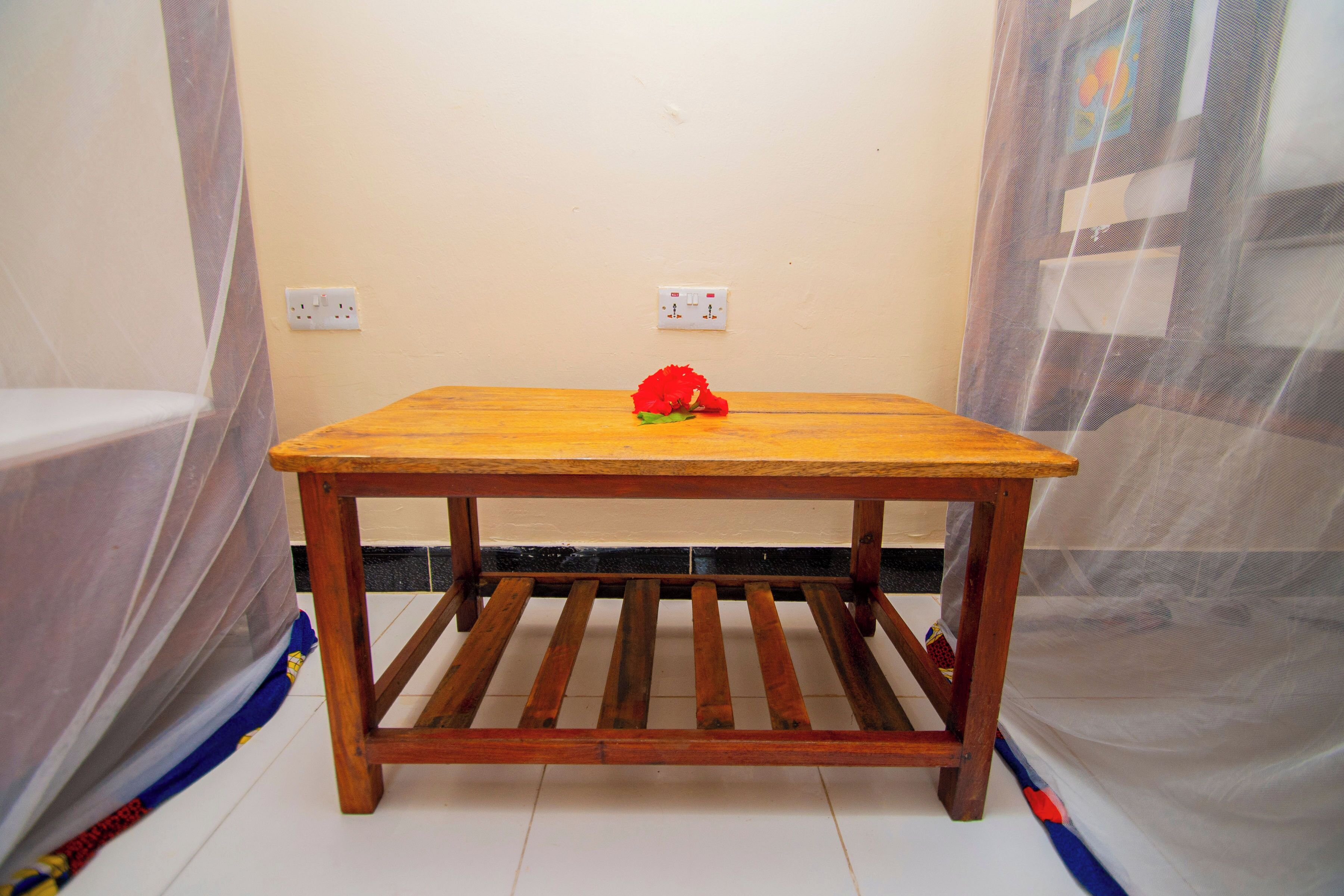 A wooden table with a red flower on it, positioned in a room with a plain wall and a power outlet.
