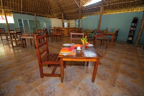 Un comedor con mesas y sillas de madera, piso de baldosas y techo de paja.