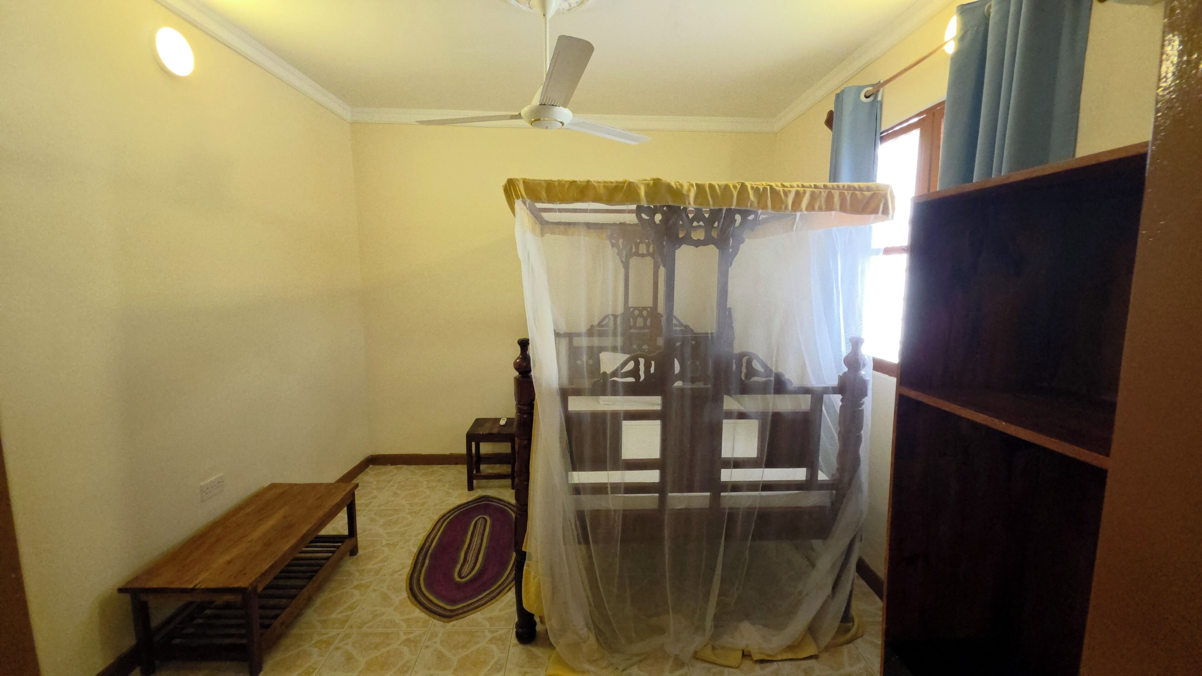 Une pièce comprenant un lit, un ventilateur de plafond, un banc et une fenêtre avec des rideaux.