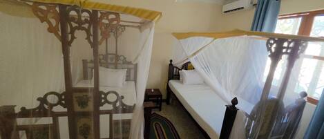 A room with two beds, one with a canopy and one without, a ceiling fan, an air conditioning unit, and a window with curtains.