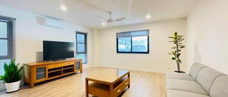 A spacious living area with a wooden coffee table, a sofa, a TV on a wooden cabinet, and a ceiling fan.