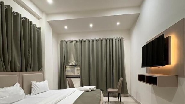 A modern hotel room with a large bed, a ceiling fan, a TV mounted on the wall, and a small seating area. - Shft-In Hotel (Noida)