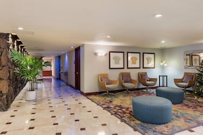 Een hotel lobby met een stenen muur, een betegelde vloer, rieten stoelen, een blauwe poef en ingelijste kunstwerken aan de muren.