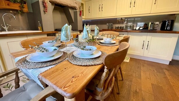 Un coin repas avec une table en bois dressée pour quatre personnes, entouré d’armoires blanches et avec une cuisine en arrière-plan.