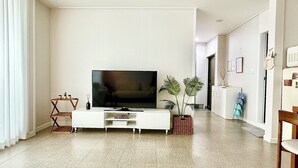 A modern living room with a flat-screen TV on a white entertainment unit, a small side table with a vase, and a potted plant.