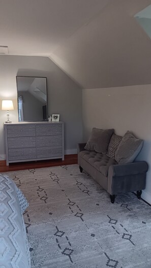Ein Schlafzimmer mit grauem Sofa, einem Schminktisch, einem Spiegel und einem gemusterten Teppich.