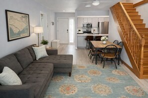 A living room with a sectional sofa, a dining table with chairs, a kitchen area with appliances, and a wooden staircase. - Resort Club Cottage on the lagoon (Edisto Island)