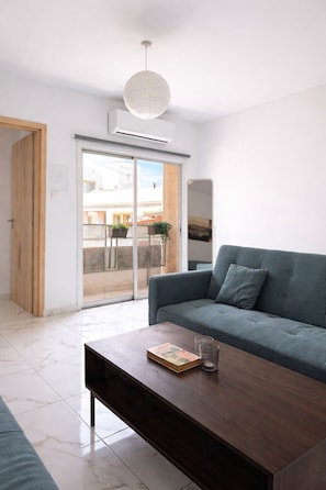 A modern living room with a grey sofa, a wooden coffee table, and a large window with a view of buildings. - Cozy 2BR - 1Min To The Beach (Larnaka)