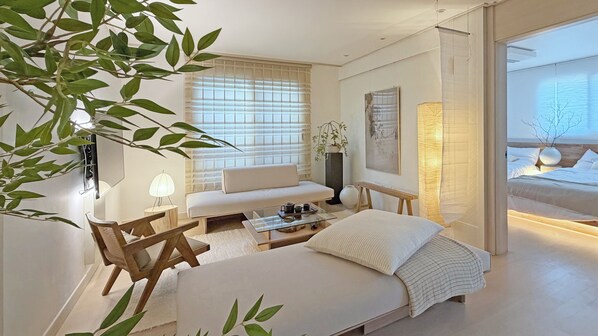 A modern, minimalist living room with a sofa, a coffee table, and a bed. There is a large window with blinds, a potted plant, and a wall-mounted lamp.