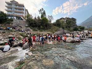 A group of people near a river with large rocks, a building with multiple floors, and a mountainous backdrop. - Manuni Inn  Khanyara Rd (Dharamshala)