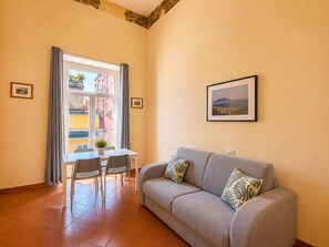 Una sala de estar con un sofá, una mesa de comedor con sillas, una ventana con cortinas y un cuadro enmarcado en la pared.