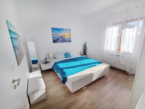 Een slaapkamer met een bed, nachtkastje, lamp, raam met gordijnen en een schilderij aan de muur.