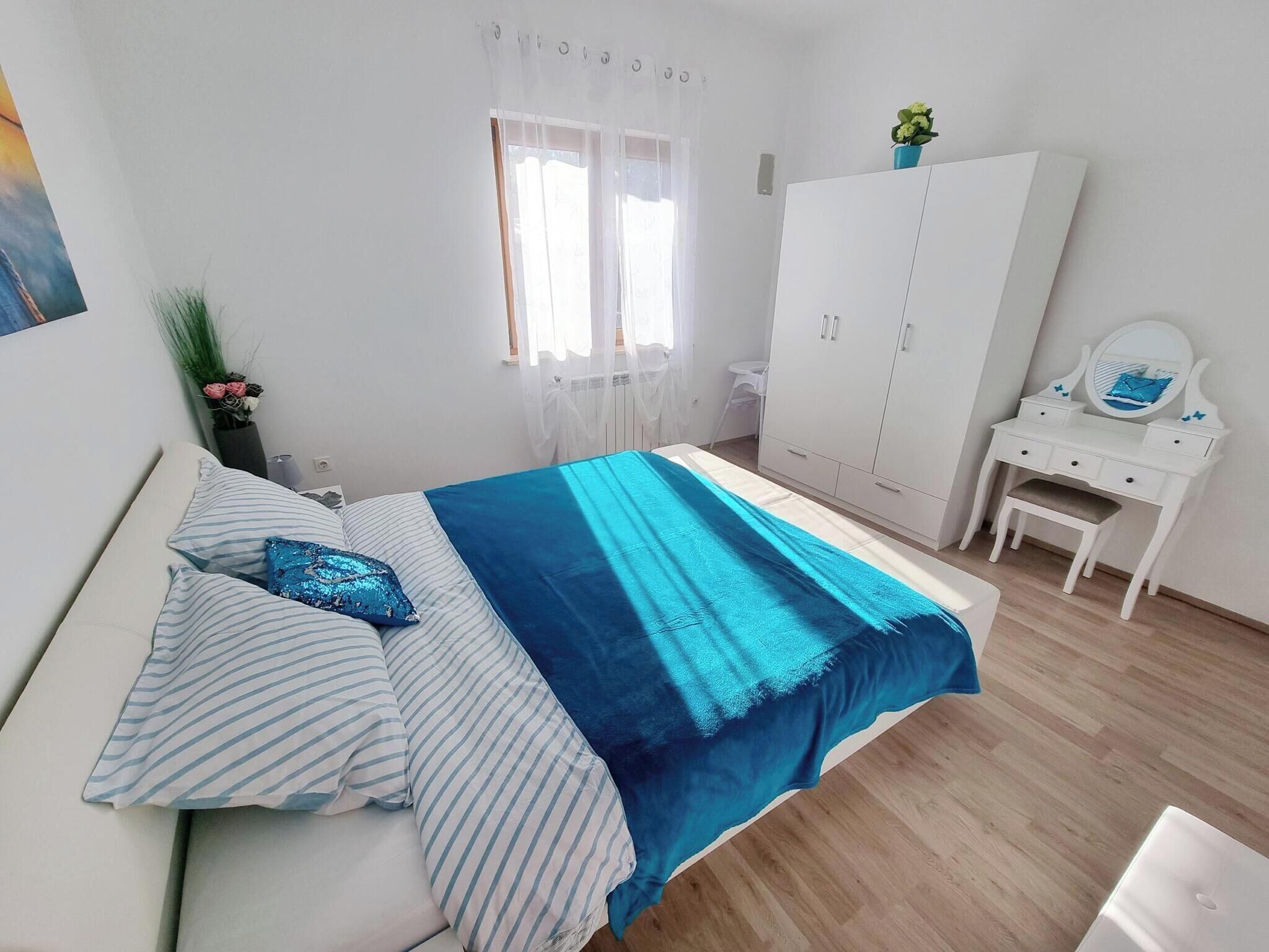 Ein Schlafzimmer mit einem Bett, einem weißen Kleiderschrank, einem Frisiertisch mit Spiegel und einem Fenster mit durchsichtigen Vorhängen.