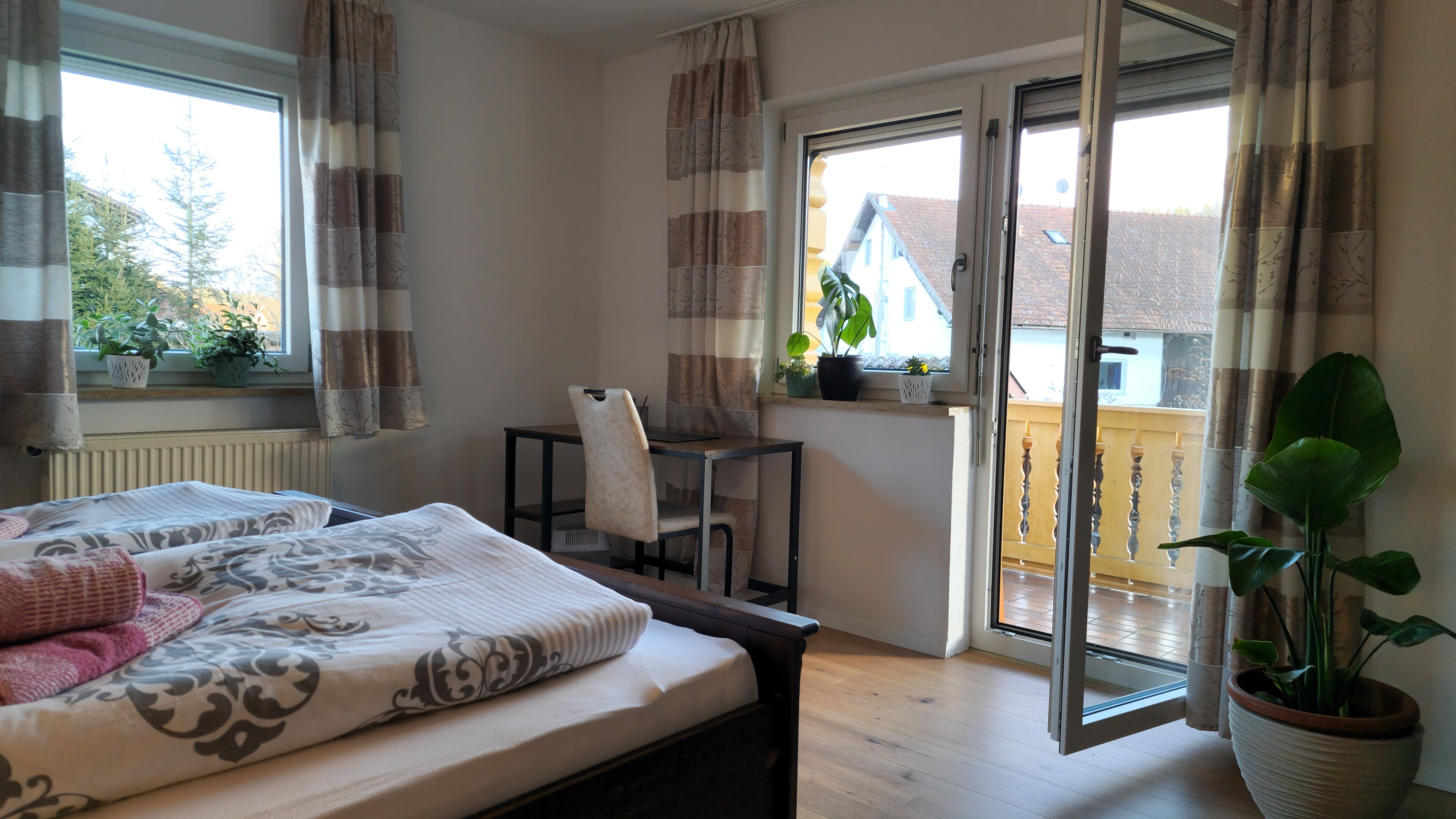 Une chambre à coucher comprenant un lit, un bureau, une chaise, une plante en pot et une porte-fenêtre coulissante donnant sur un balcon extérieur.