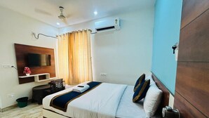 A hotel room with a bed, a TV mounted on the wall, an air conditioning unit, a ceiling fan, and a wooden door. - S K Comforts Rooms (Bengaluru)