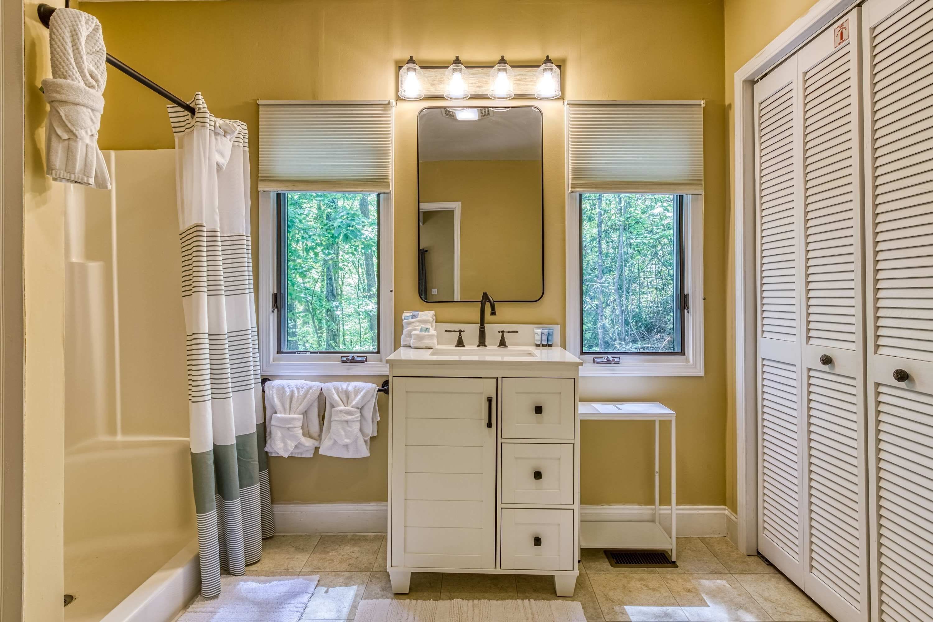 A bathroom with a bathtub, a vanity with a mirror and lights, a window with blinds, and a towel rack.