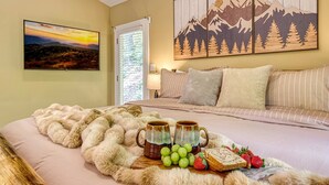 Een slaapkamer met een bed, een schilderij van bergen, een raam met jaloezieën en een dienblad met fruit en mokken.
