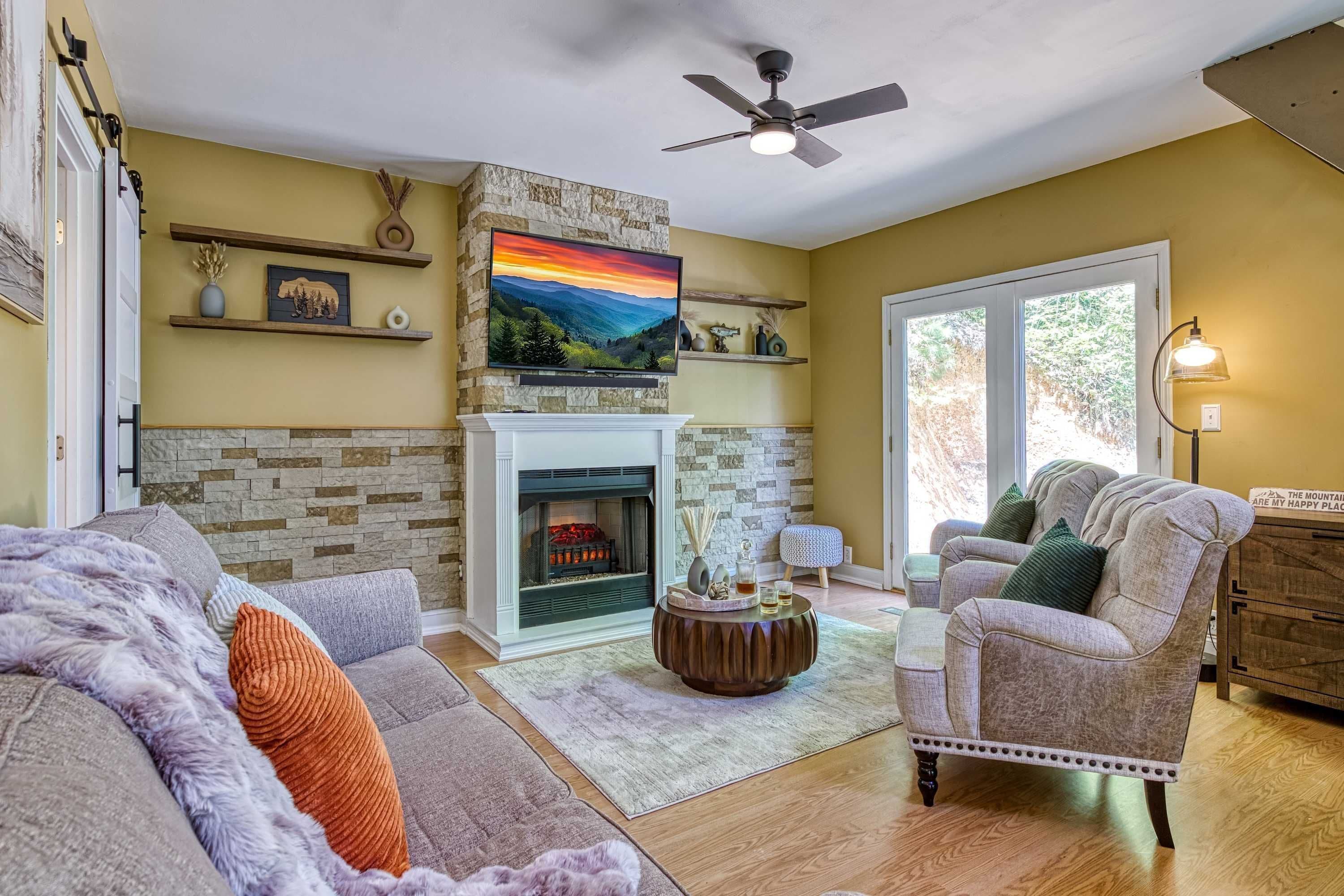 A living room with a fireplace, a ceiling fan, and a large window.