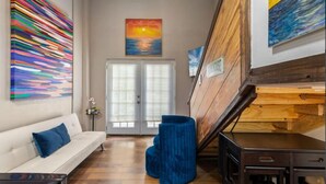 Un intérieur moderne avec un canapé blanc, un pouf bleu, un escalier et un tableau représentant un coucher de soleil.