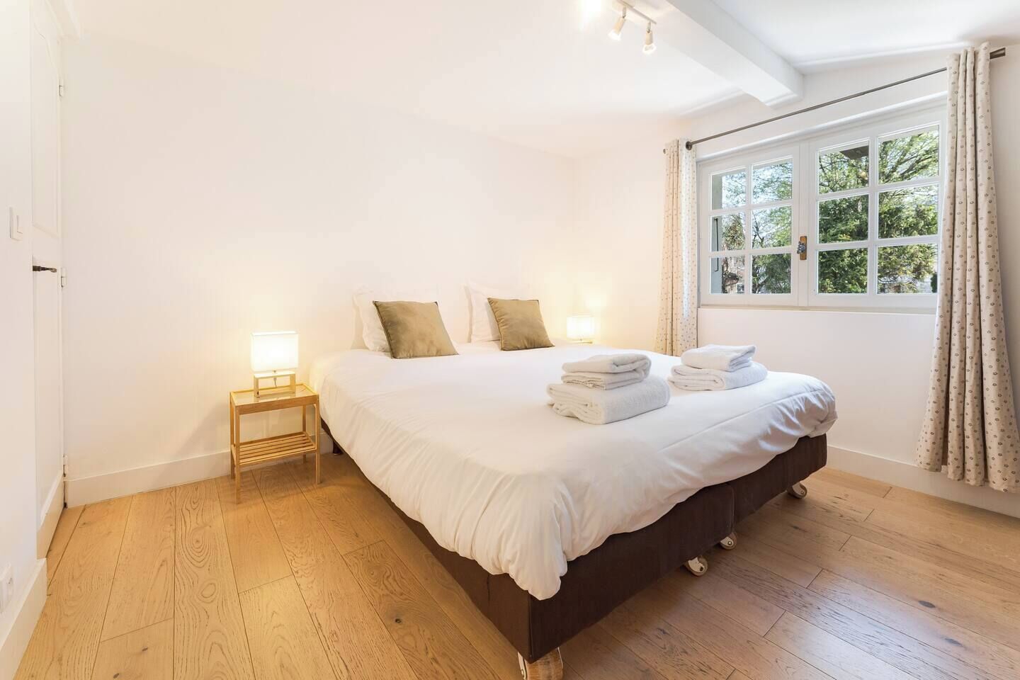 A bedroom with a large bed, a bedside table with a lamp, and a window with curtains.