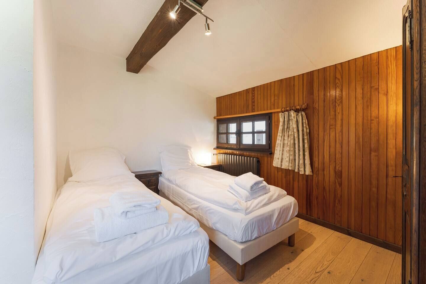 A bedroom with two beds, wooden paneling, a window, and a radiator.