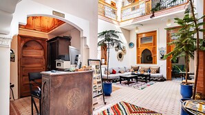 A spacious living area with a sofa, a coffee table, and a patterned rug. There is a wooden desk with a computer and a chair. The room has a high ceiling with a balcony.
