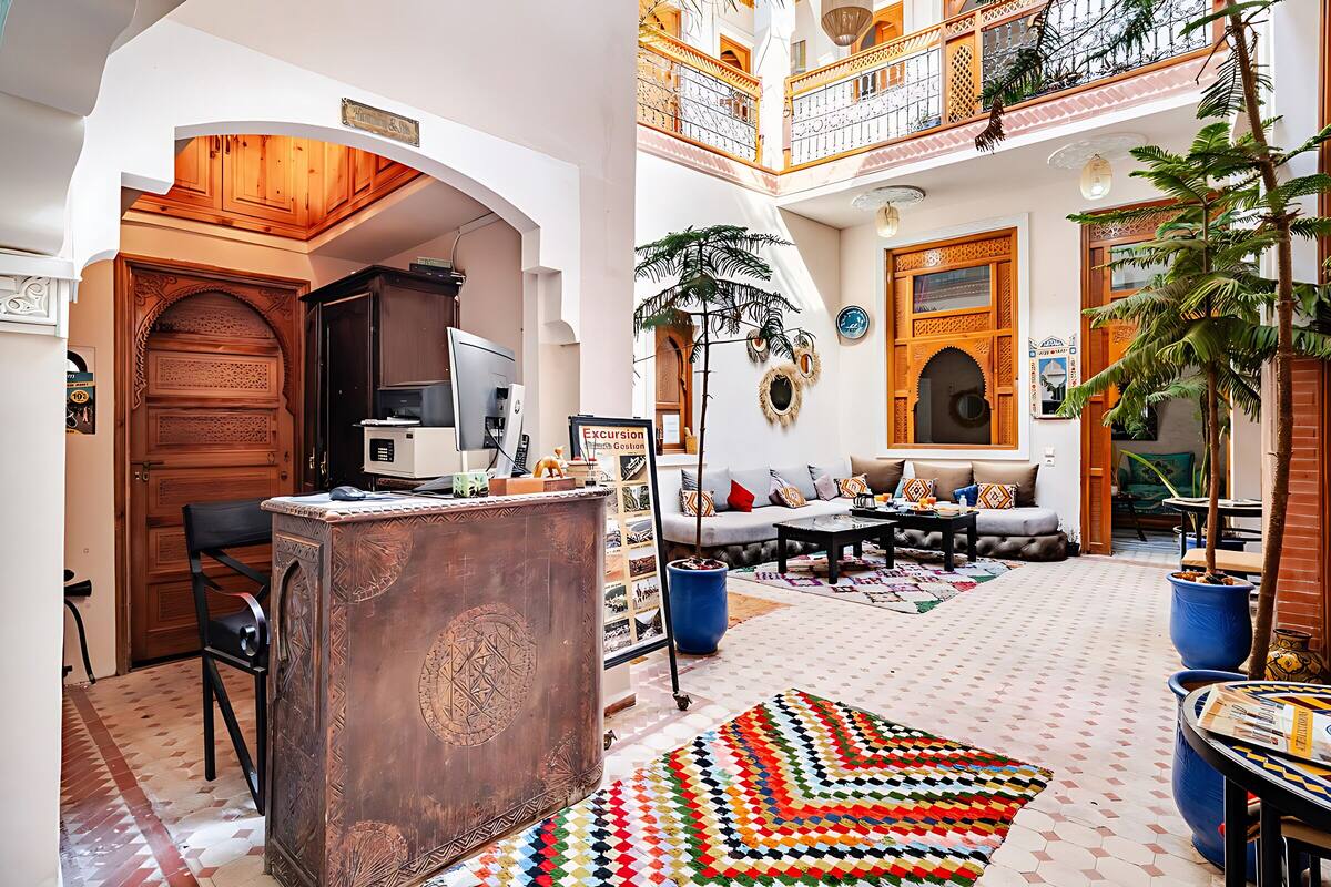 A spacious living area with a sofa, a coffee table, and a patterned rug. There is a wooden desk with a computer and a chair. The room has a high ceiling with a balcony.